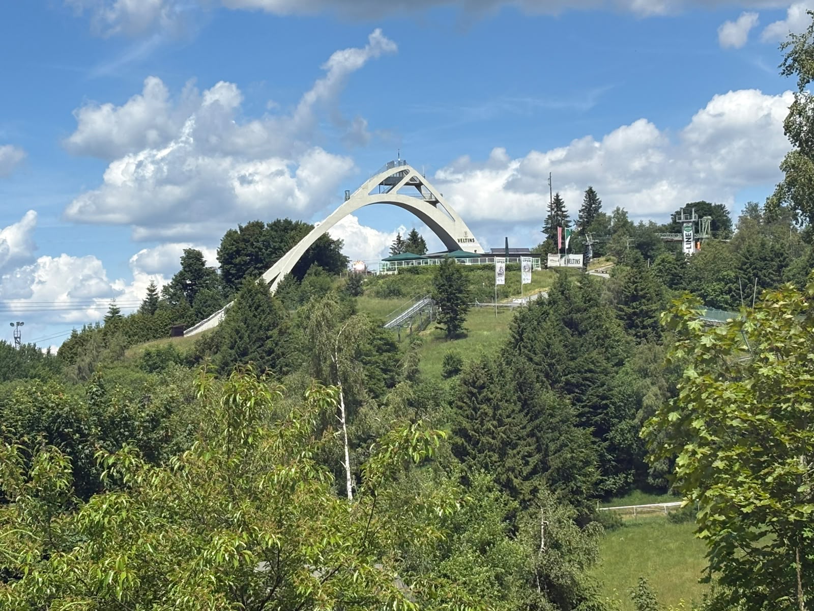 Blick von der Astenstraße auf die Sprungschanze, eingebettet in die grüne Landschaft und vor blauem Himmel